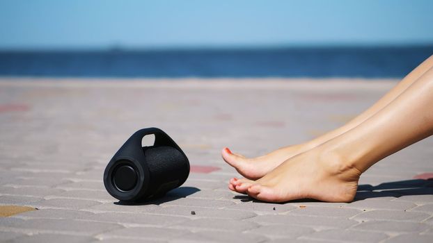 close-up, mini music bluetooth portable black cylinder wireless loudspeaker. female legs in silver sandals, with bright red pedicure, dancing to the music. on the beach, in summer. High quality photo