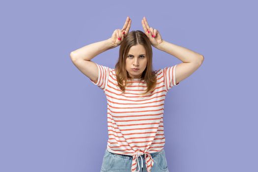 Portrait of angry bossy blond woman wearing striped T-shirt standing and showing horns, being anger and aggressive, looking at camera. Indoor studio shot isolated on purple background.
