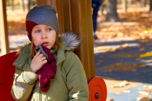 a young human being below the age of puberty or below the legal age of majority. Offended frightened child in a jacket and a hat on an autumn playground.