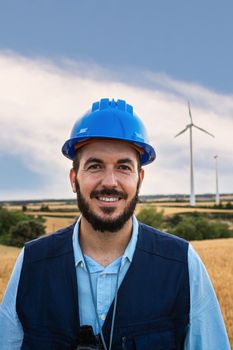 Vertical portrait of happy electric engineer standing at wind turbine farm looking at camera. Renewable energy concept.