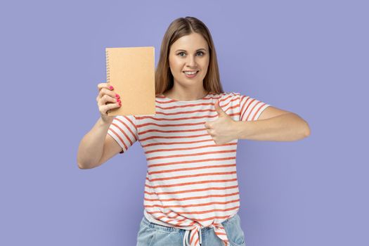 Portrait of delighted smiling blond woman wearing striped T-shirt holding book, looking at camera and showing thumb up, recommend novel for reading. Indoor studio shot isolated on purple background.