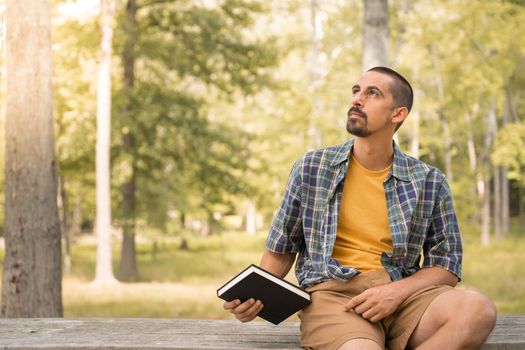 Young man sitting outdoors holding book in a park with trees background - Knowledge faith concept. High quality photo