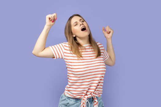 Portrait of tired blond woman wearing striped T-shirt standing yawning with closed eyes, stretching arms, feeling exhausted need rest. Indoor studio shot isolated on purple background.