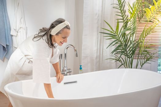 Happy smiling beautiful middle aged woman in bathrobe preparing bathtub. Bodycare spa procedures antiage recreation skin care and beauty salon concept.