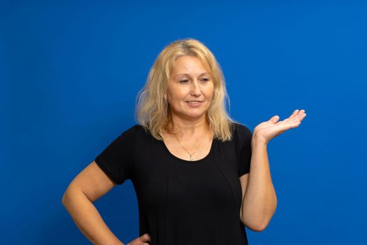 Beauty offer. Smiling attractive middle age woman holding something on her empty palm, happy woman with beautiful skin announcing new cosmetic. Isolated on blue studio background.