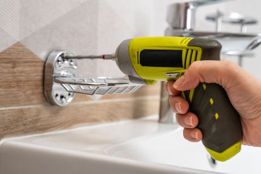 a close-up of an electric cordless screwdriver that wraps a screw into a hole in a ceramic tile to install a soap dish and shelves. home renovation. installing and fixing bathroom accessories