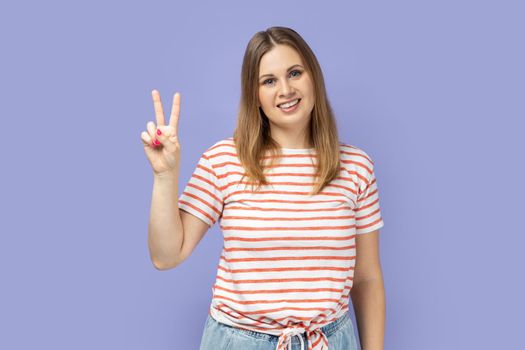 Portrait of beautiful blond woman wearing striped T-shirt smiling happily and showing v sign or peace gesture, rejoicing win. Indoor studio shot isolated on purple background.