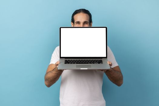 Portrait of man with beard wearing white T-shirt covering half of face behind laptop with white empty display, looking at camera with kind eyes. Indoor studio shot isolated on blue background.