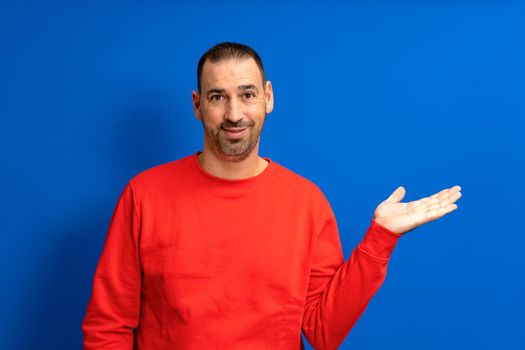 Bearded hispanic man in red sweater over blue background smiling kindly offering something with open hand or as greeting and welcome. Successful business.