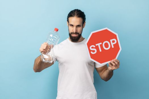 Portrait of strict responsible man with beard wearing white T-shirt holding plastic bottle and stop red sign, worrying about environment. Indoor studio shot isolated on blue background.