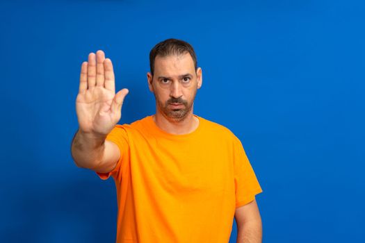 Bearded hispanic man dressed in orange t-shirt making stop gesture with palm of hand isolated on blue studio background. He is fed up with the social and labor injustices caused by the current system