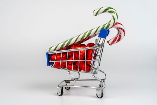 A supermarket trolley loaded with heart-shaped candies and Christmas caramel canes on a white background. Gifts and purchases for the New Year and Christmas. Online trading. A toy shopping cart carries a Christmas candy cane