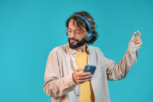 Happy attractive man dancing on blue background with headphones and holding smartphone. High quality photo