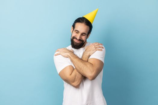 Portrait of smiling handsome birthday man with beard wearing white T-shirt and party cone, celebrating holiday, hugging himself, egoist. Indoor studio shot isolated on blue background.