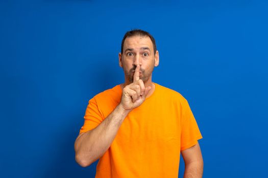 Bearded latino man wearing orange casual t-shirt, pressing finger to lips as if asking to keep quiet, isolated over blue background. shhh gesture