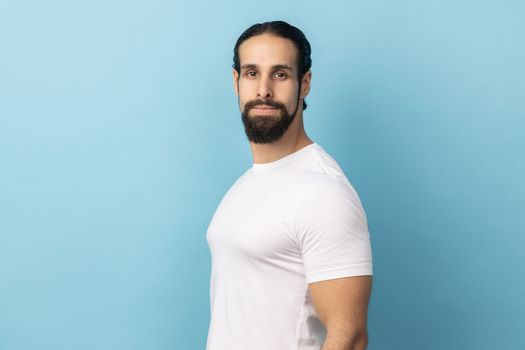 Side view portrait of bearded handsome man wearing white T-shirt standing looking at camera with serious face, being strict and bossy. Indoor studio shot isolated on blue background.