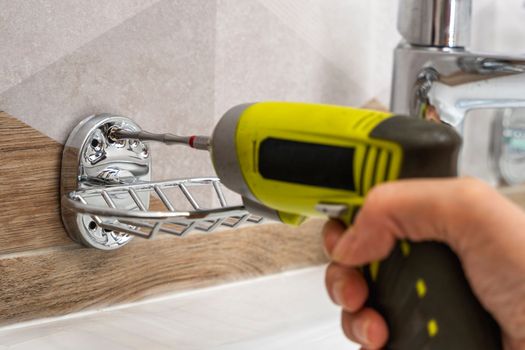 a close-up of an electric cordless screwdriver that wraps a screw into a hole in a ceramic tile to install a soap dish and shelves. home renovation. installing and fixing bathroom accessories