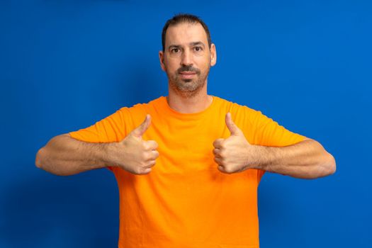 Attractive cheerful fit bearded latino man wearing orange t-shirt standing isolated over blue background, showing thumbs up.