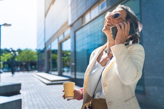 Attractive middle aged business woman with big smile talking with her smart phone with coffee cup in the street. High quality photo