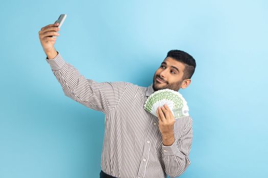 Rich businessman holding money and taking selfie by mobile phone, smiling boasting of big profit, successful online betting, wearing striped shirt. Indoor studio shot isolated on blue background.