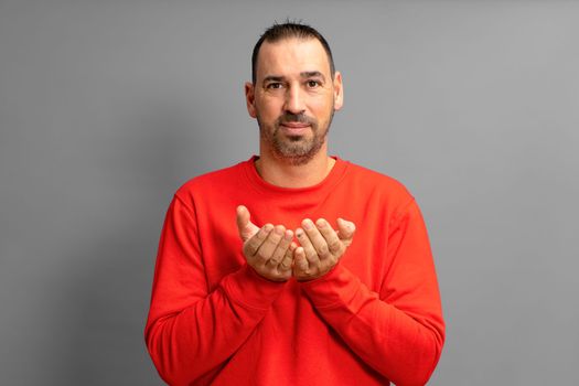 Latino man with beard dressed in a red jumper offering something imaginary in his hands close to the chest isolated on gray studio background, advertising concept, copy space.