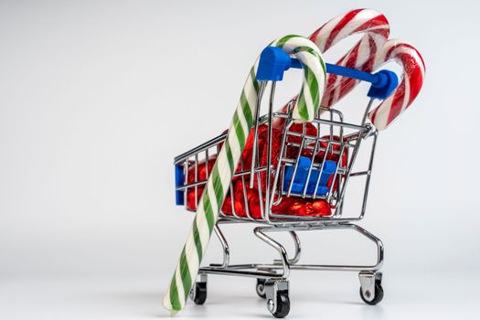 A supermarket trolley loaded with heart-shaped candies and Christmas caramel canes on a white background. Gifts and purchases for the New Year and Christmas. Online trading. A toy shopping cart carries a Christmas candy cane