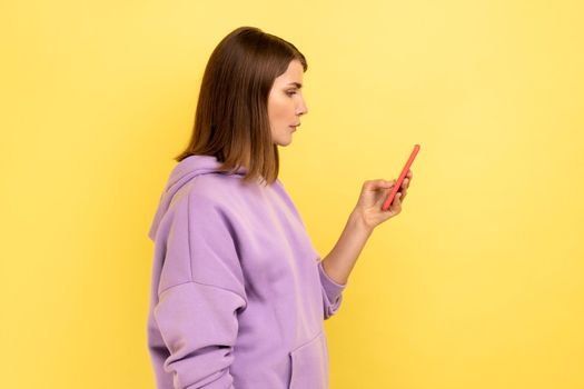 Side view of calm serious woman reading message on smartphone, using mobile device for communication, browsing web, wearing purple hoodie. Indoor studio shot isolated on yellow background.