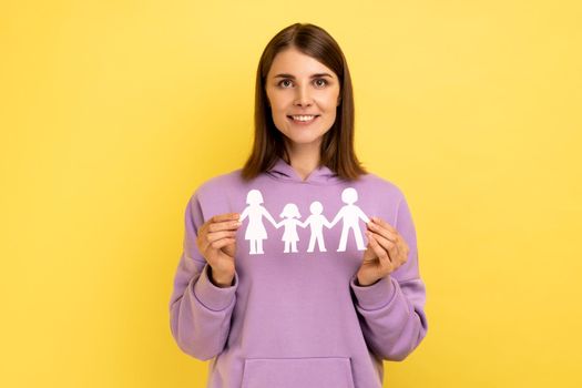 Kind smiling positive woman holding paper chain people which holding in hands, happy family, relationships, parenthood, wearing purple hoodie. Indoor studio shot isolated on yellow background.