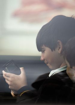 Close up portrait of kids with school uniform through the car window glass playing with smartphone. Side view.