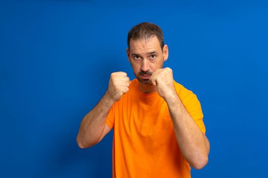 Caucasian man wearing orange t-shirt over blue background ready to fight with fist defense gesture, angry and upset face, fear of problem.
