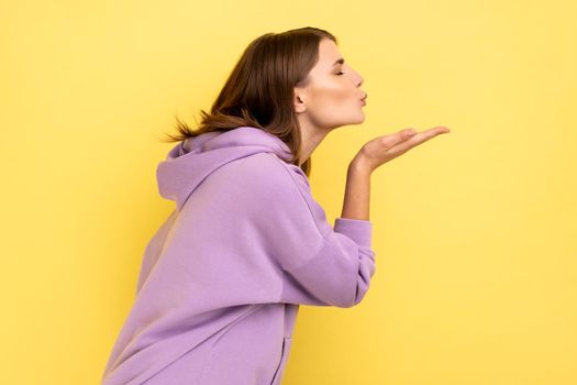 Side view of woman sending air kiss, flirting, demonstrating love affection feelings, keeps eyes closed, romantic relationships, wearing purple hoodie. Indoor studio shot isolated on yellow background