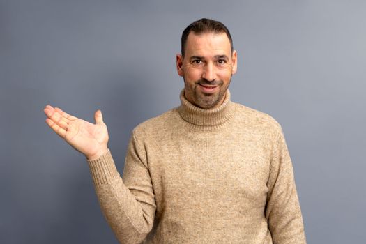 Hispanic man with beard and brown turtleneck sweater in friendly attitude offering something on the palm of his hand, isolated on gray background