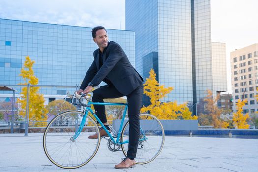 Young smiling stylish African American businessman in suit going to the office by bicycle in front of financial district. Business and transport concept. High quality vertical photo