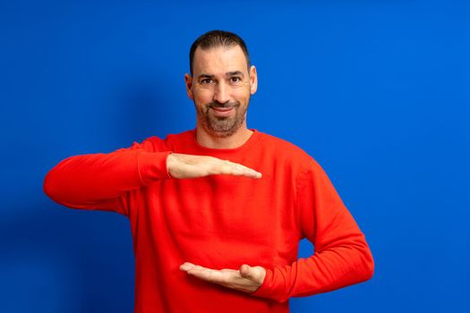 A man holding an object with both hands in front of him, displaying, offering or advertising an object against a blue wall. Empty. Space. Concept. Object. Background. Portrait. Copy.