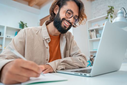 Happy young creative entrepreneur laughing smiling writes in a notebook next to laptop in a modern workspace office at home. Freelance guy starting small business. High quality photo