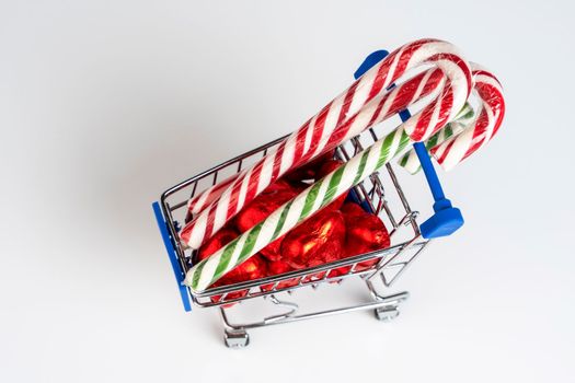 A supermarket trolley loaded with heart-shaped candies and Christmas caramel canes on a white background. Gifts and purchases for the New Year and Christmas. Online trading. A toy shopping cart carries a Christmas candy cane