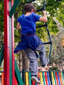 Rear view of child boy climbing on the rope ladder in a park playground.