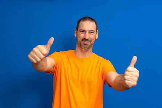 Portrait of cheerful man in basic clothing smiling and showing thumbs up at camera isolated over blue background