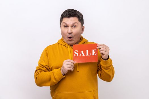 Portrait of astonished man holding sale card in hands looking at camera with open mouth, black Friday, wearing urban style hoodie. Indoor studio shot isolated on white background.