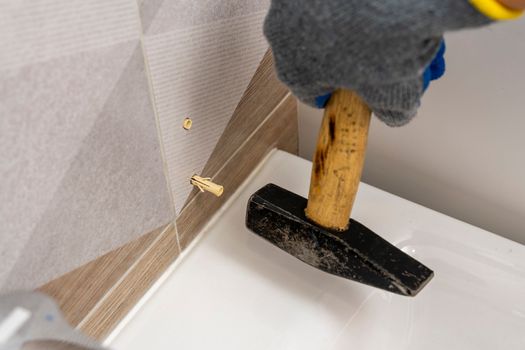 A worker's hand in protective gloves hammers a plastic dowel pin into a drilled hole in the wall against the background of the bathroom interior. renovation in the bathroom. installation of bathroom accessories.