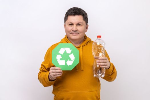 Portrait of delighted dark haired man with smile holding empty plastic bottle and green recycling sign, wearing urban style hoodie. Indoor studio shot isolated on white background.