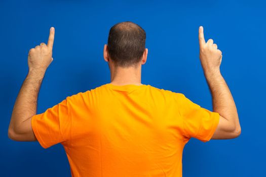 Large man with receding hairline on his back pointing up with his index fingers isolated on blue studio background