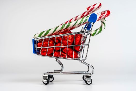 A supermarket trolley loaded with heart-shaped candies and Christmas caramel canes on a white background. Gifts and purchases for the New Year and Christmas. Online trading. A toy shopping cart carries a Christmas candy cane