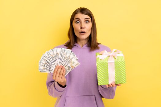 Portrait of excited amazed woman holding wrapped present box and doolars banknotes, looking at camera with open mouth, wearing purple hoodie. Indoor studio shot isolated on yellow background.