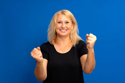 Closeup of emotional attractive blonde caucasian woman in black t-shirt with hands in fists, isolated on blue background