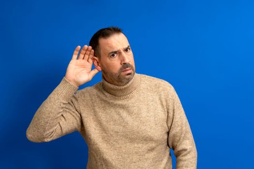 Bearded latino man wearing turtleneck listening holds his hand near his ear on a blue background. Handsome man trying to listen to someone's conversation.