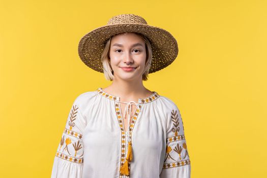 Portrait of young ukrainian blonde woman in traditional embroidered blouse on yellow studio background. Confident sunny outfit, straw hat. High quality photo