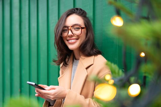 Woman using smartphone on Christmas market. Caucasian female enjoying winter holiday season. Concept of social networking, communicating, using app. Blurred Christmas lights on foreground. Holiday