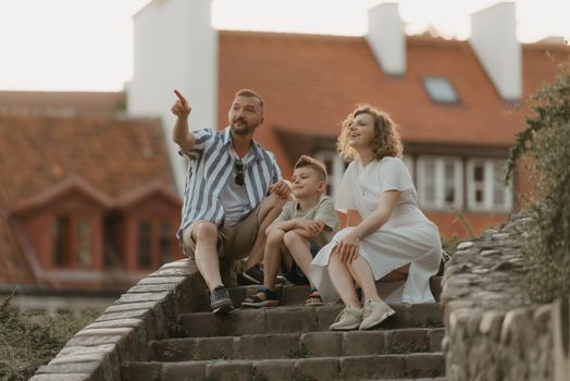 A smiling family is staring to the side on the stairs between roofs in an old European town. A happy father, mother, and son are having fun in the evening.