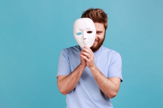 Portrait of positive bearded man removing white mask from face showing his smiling expression, good mood, pretending to be another person. Indoor studio shot isolated on blue background.